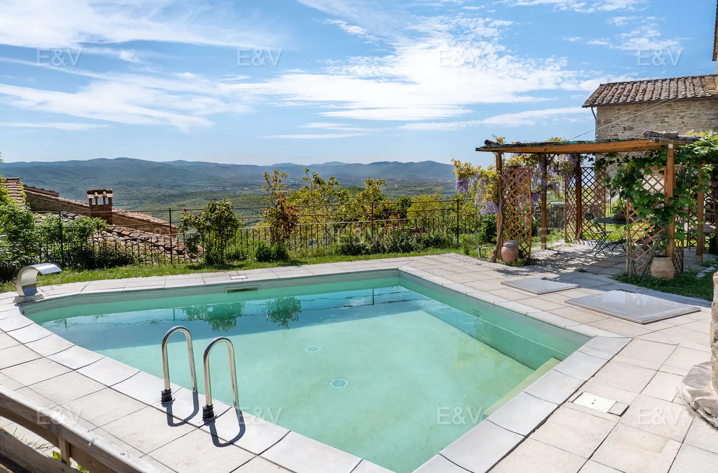Outdoor pool with metal ladder, surrounded by stone patio and greenery, overlooking a valley under a blue sky.