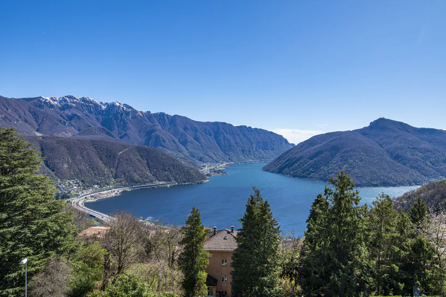 Scenic view of Lake Lugano, Switzerland, with blue water, green trees, and snow-capped mountains under a clear blue sky.