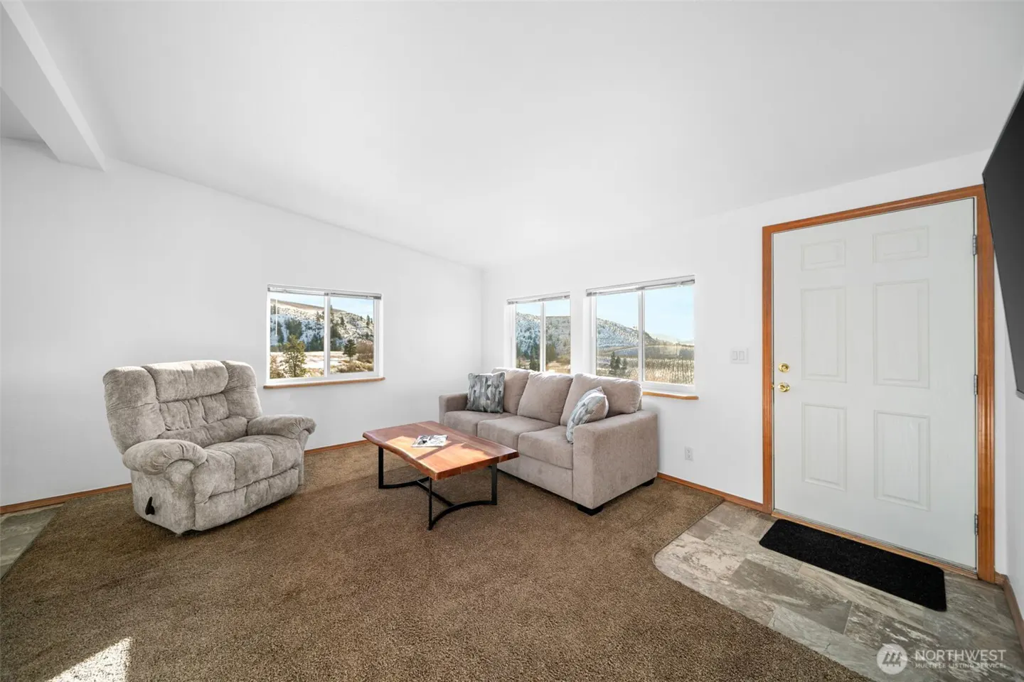 Living room with white walls, brown carpet, beige sofa, recliner, and wood coffee table. Windows overlook a snowy landscape.