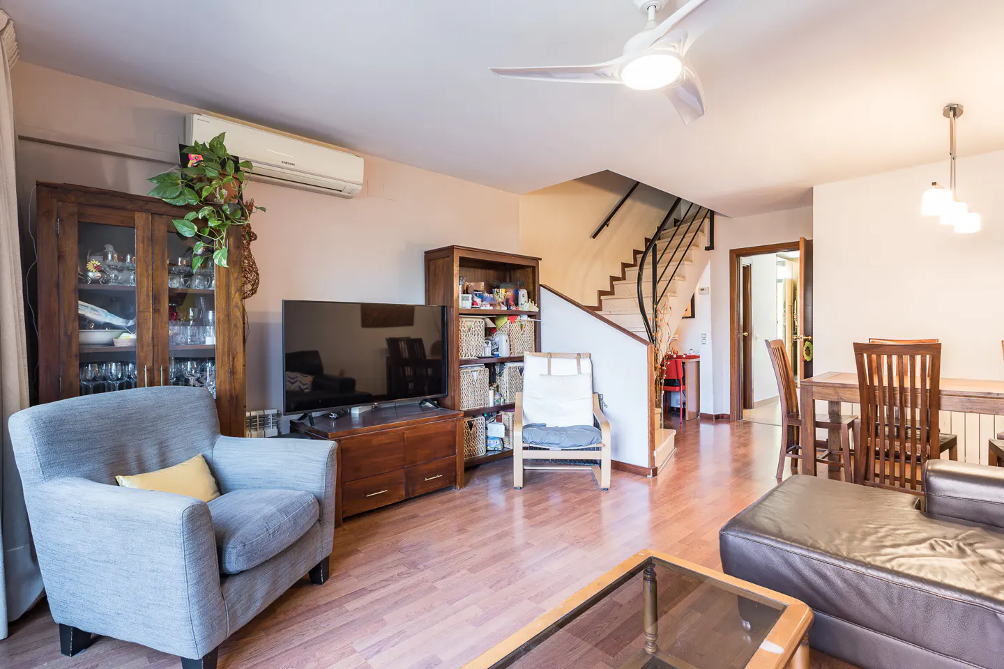 Living room with wood floors, gray armchair, TV, and staircase. A ceiling fan and dining area are also visible.