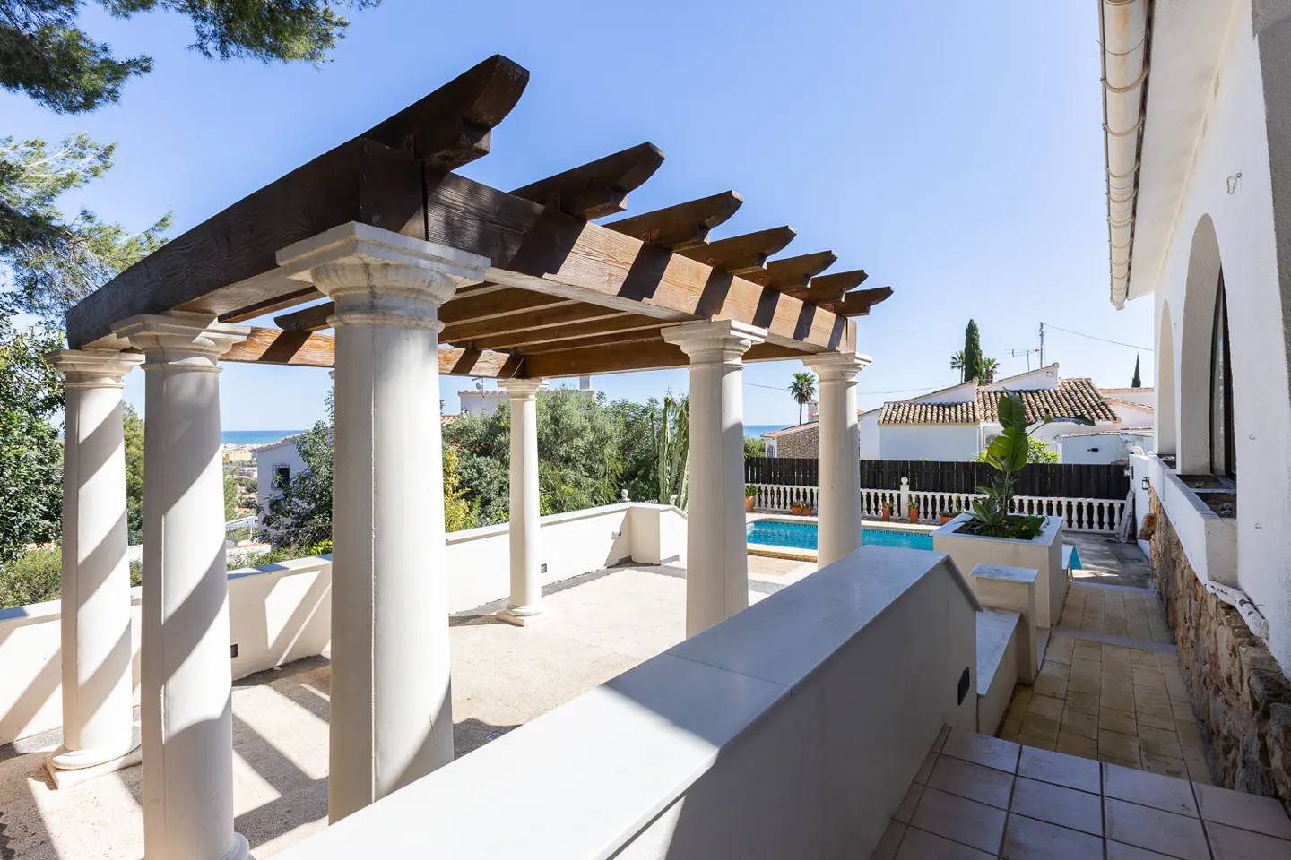 Outdoor patio with white columns and a wooden pergola overlooking a pool and neighborhood on a sunny day.