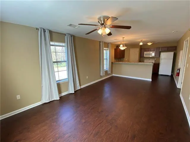 Open-concept living space with dark wood floors, tan walls, and a ceiling fan. Kitchen with brown cabinets and white appliances in the background.