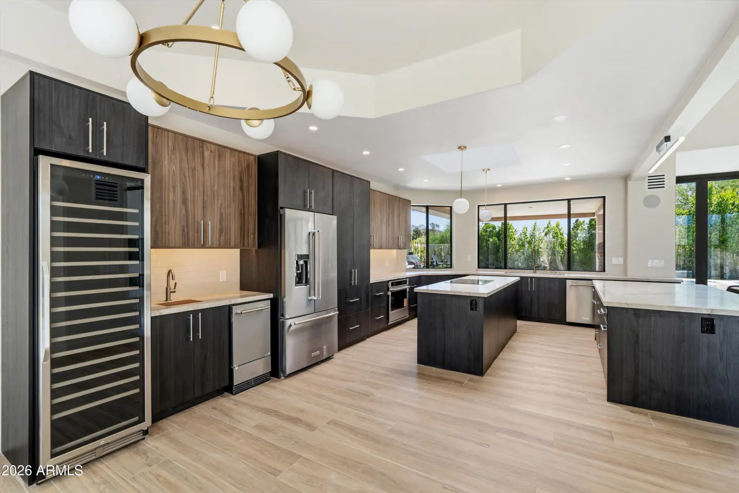 A modern kitchen with dark wood cabinets, stainless steel appliances, and a large island. A gold chandelier hangs above.