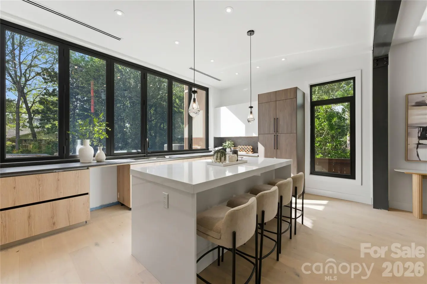 Modern kitchen with white island, beige bar stools, light wood cabinets, and large windows overlooking green trees.