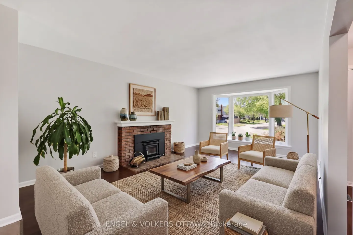 Bright living room with two beige sofas, two chairs, and a wood coffee table on a jute rug. A brick fireplace and bay window add charm.