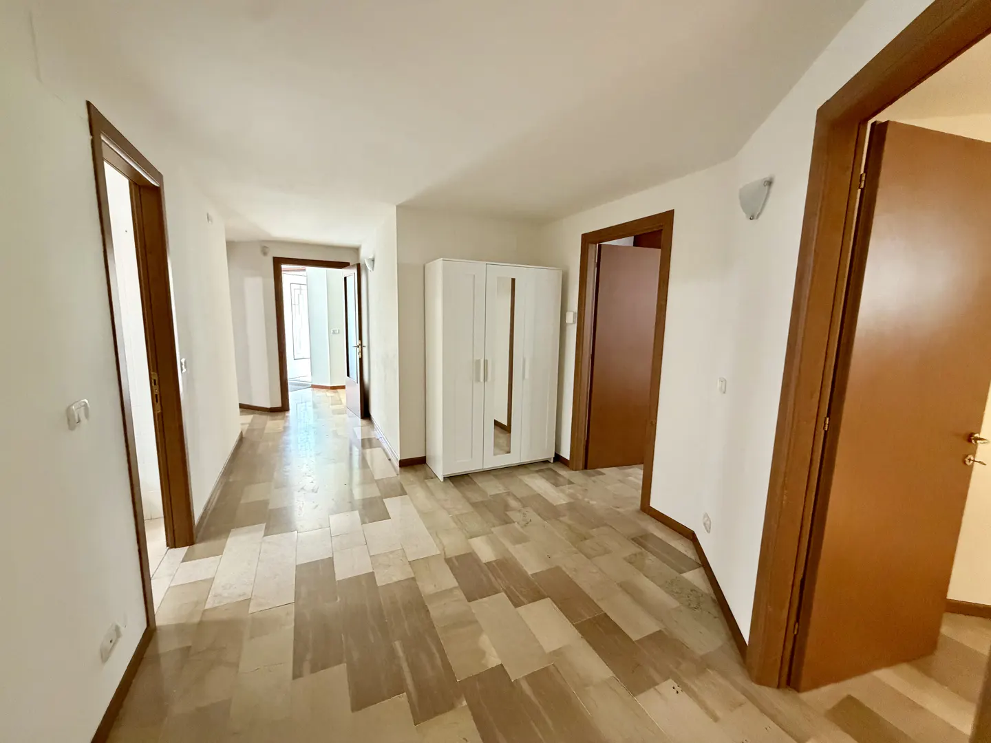 A bright hallway with white walls, brown trim, and a patterned tile floor. A white wardrobe stands near an open doorway.