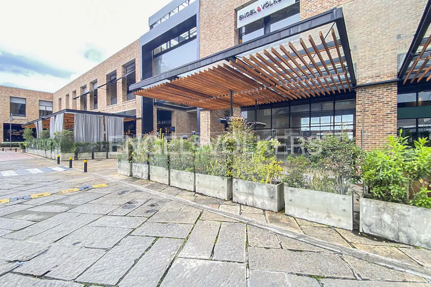 Exterior of a brick building with an Engel & Volkers sign, a wooden pergola, and concrete planters.