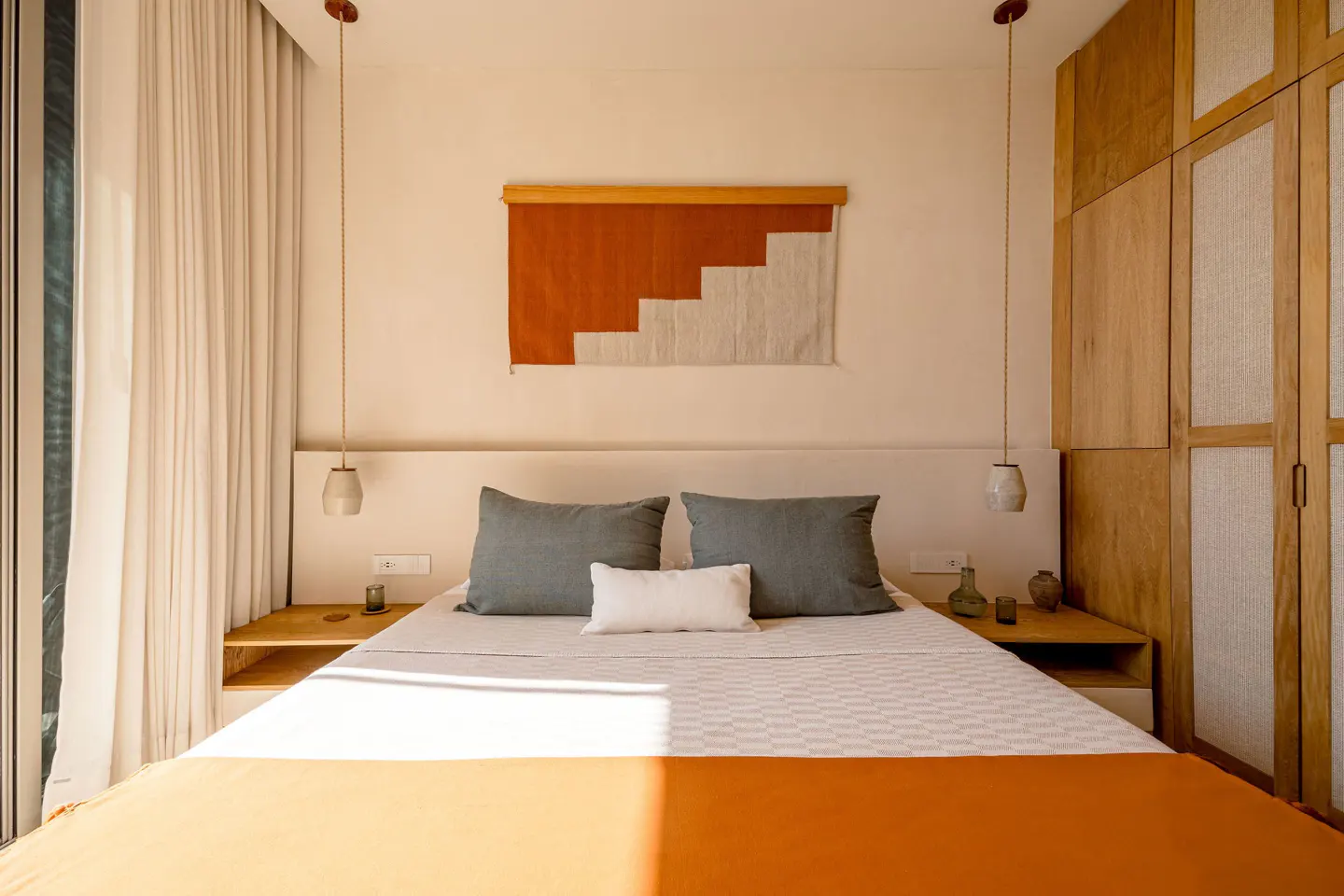 Bedroom with orange blanket, gray pillows, and white walls. A tapestry hangs above the bed. Wooden closet on the right.