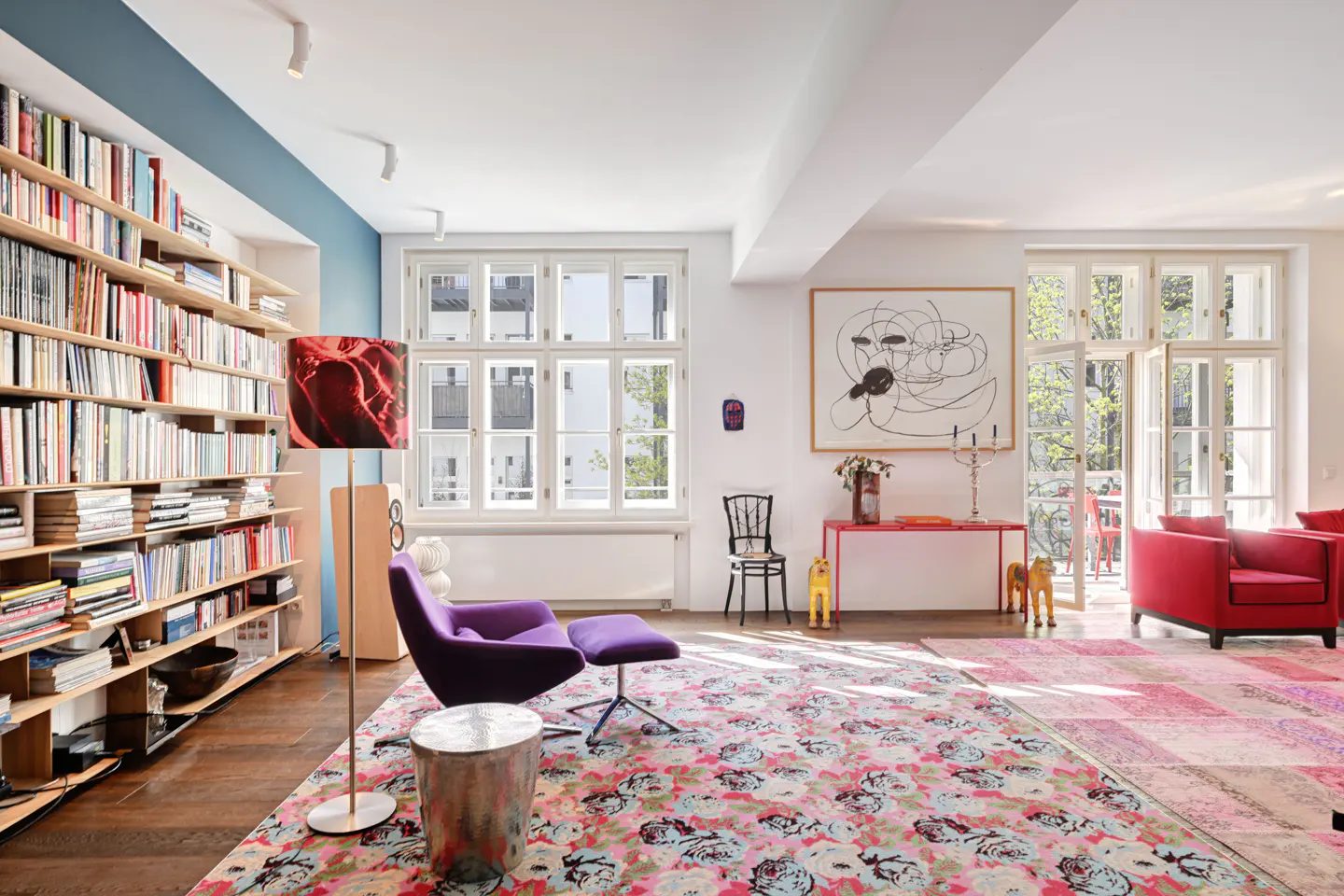 Bright living room with a large bookshelf, purple chair, and floral rug. Sunlight streams through open doors. A red table and abstract art add pops of color.