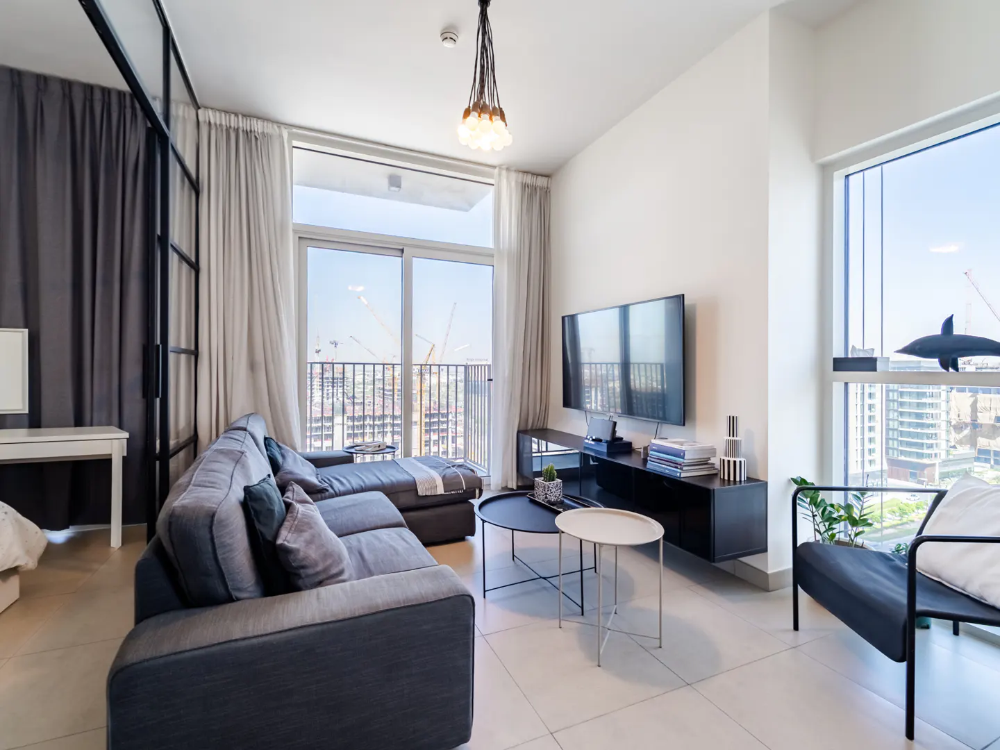 Modern living room with a grey sofa, black media console, wall-mounted TV, and floor-to-ceiling windows overlooking a city.