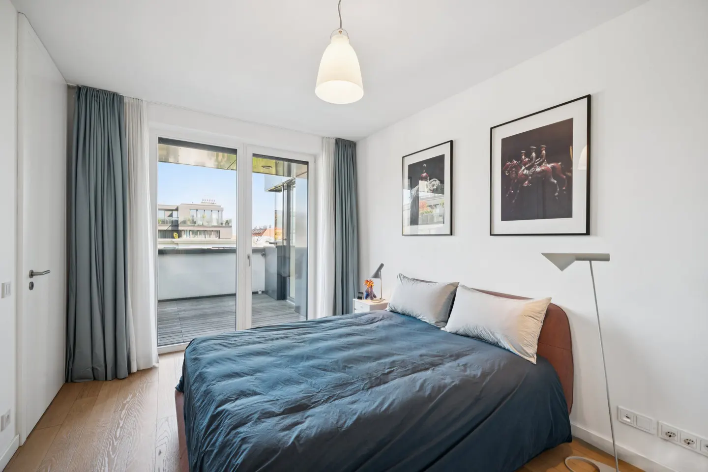 Bright bedroom with a blue duvet on the bed, white walls, and gray curtains by a glass door to a balcony.