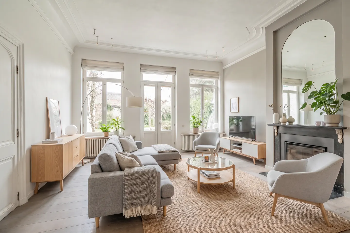 Bright, modern living room with a grey sofa, two armchairs, a round coffee table, and a fireplace with a large mirror.