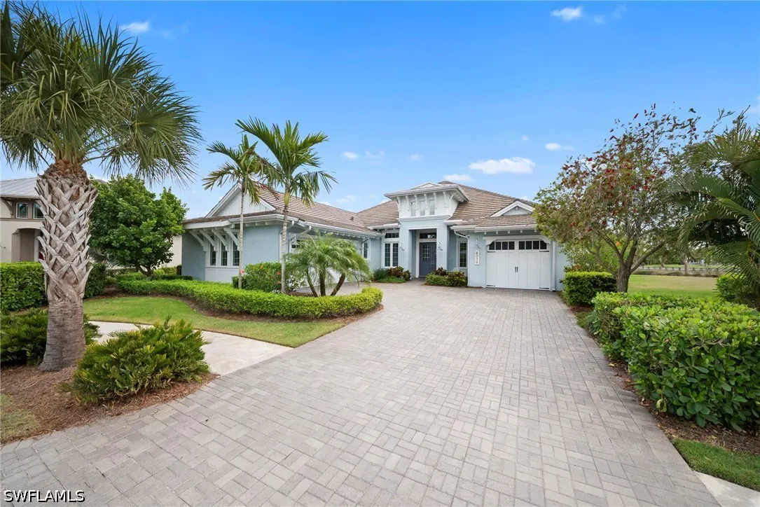Light blue single-story home with a brown roof, white trim, and a brick driveway under a clear blue sky. Palm trees and green bushes surround the house.
