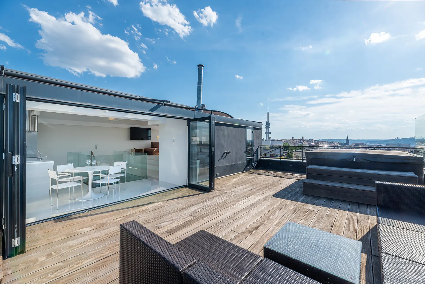 Rooftop patio with wood deck, wicker furniture, and open folding doors to a white dining area. City skyline in the background.