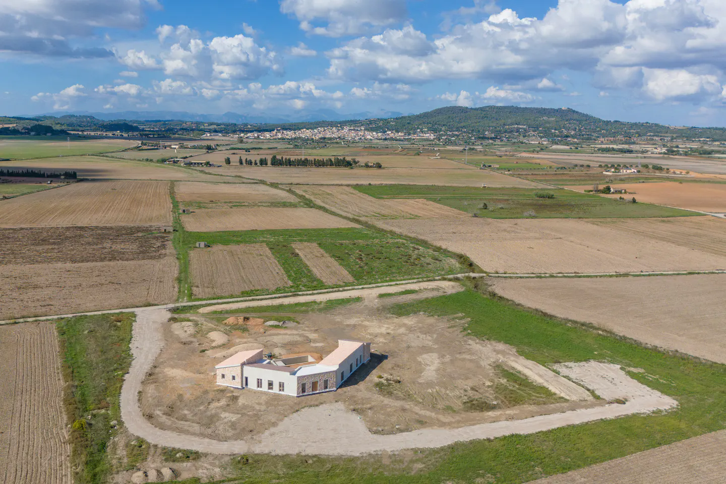 Aerial view of a modern, white, U-shaped house surrounded by fields under a blue sky with scattered clouds.
