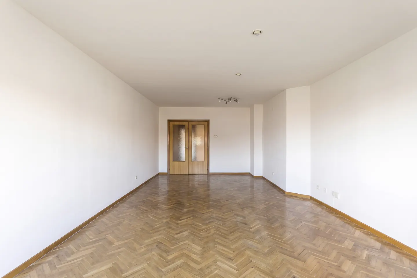 Empty room with white walls, herringbone wood floor, and double doors at the far end.