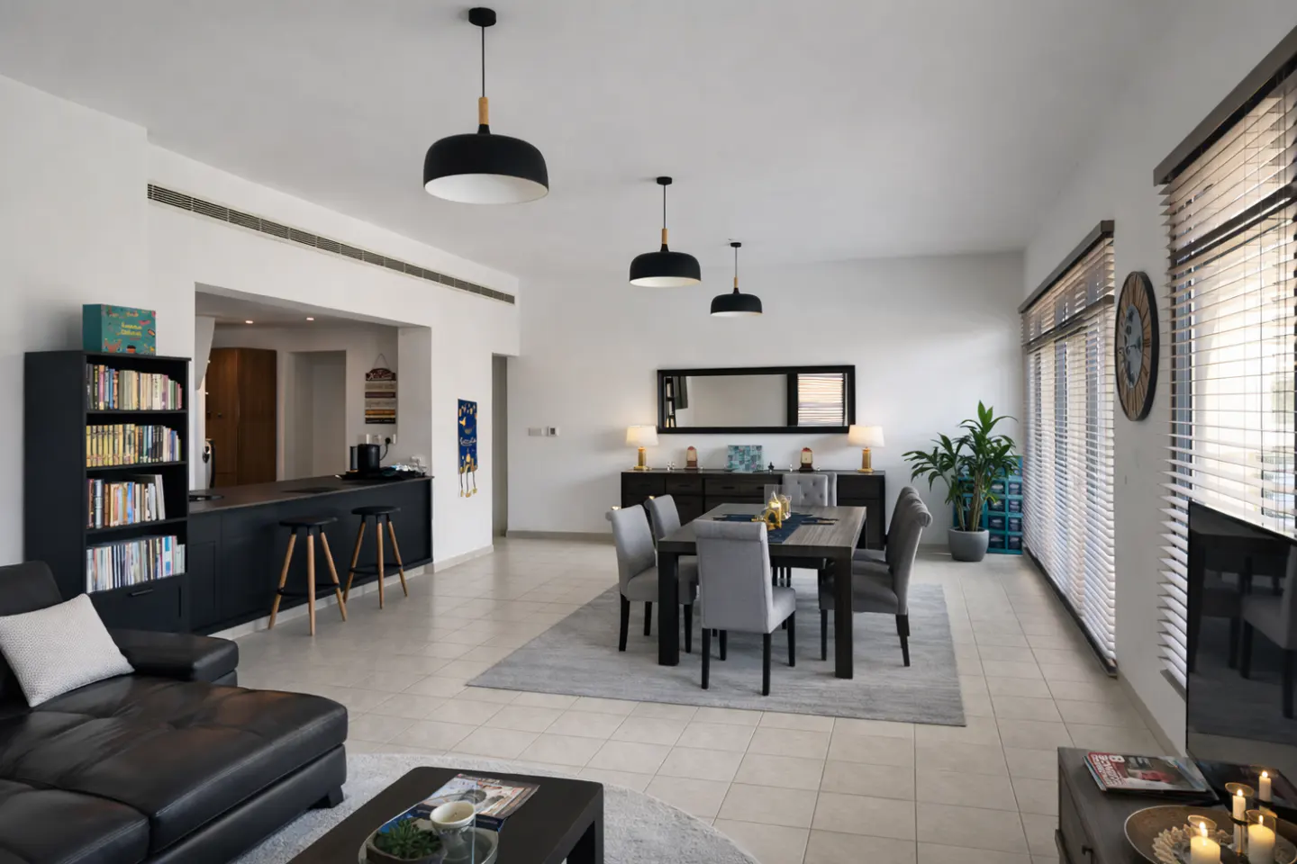 Open-concept living and dining area with black leather sofa, dining table with gray chairs, and black pendant lights.