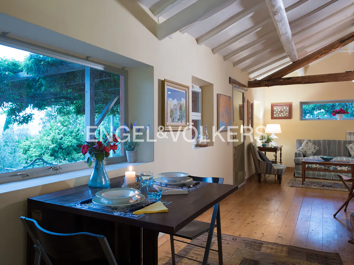 Interior view of a dining table set for two, with a view of a lush garden through a window. Living room with sofa in the background.