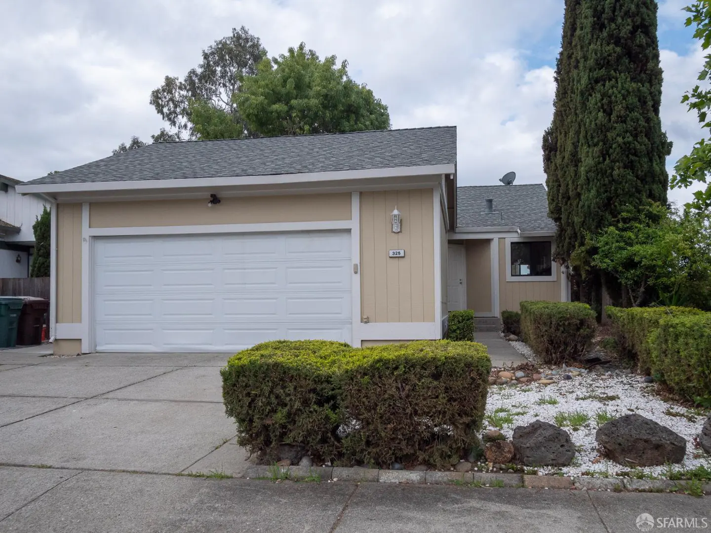 Tan single-story home with a white garage door, gray roof, and green bushes in front. A tall, thin tree stands to the right of the front door.