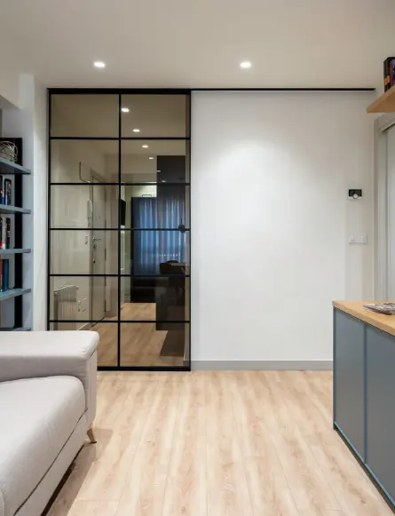 Modern living room with light wood floors, white walls, and a black-framed glass sliding door. A gray sofa and blue cabinets add color.