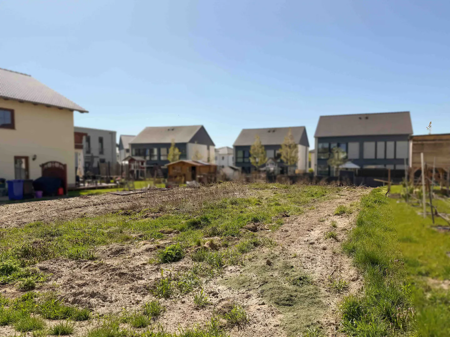 Vacant lot with dirt path and sparse grass, surrounded by modern houses under a clear blue sky.
