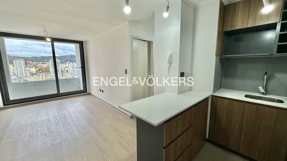 A bright apartment interior with wood floors, a kitchen island, and a large window overlooking a city.