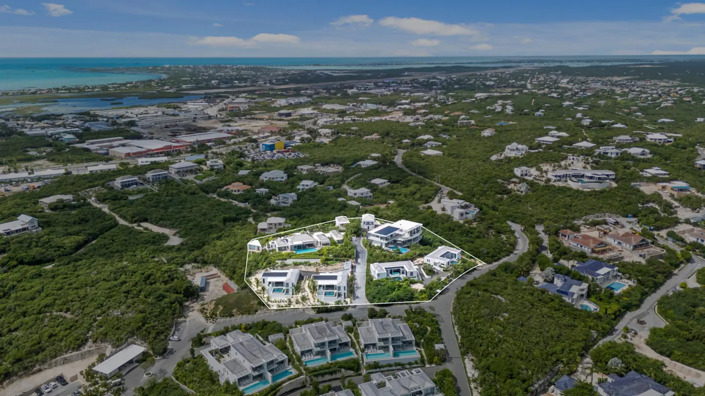 Aerial view of luxury homes with white walls and pools, surrounded by green vegetation and a blue ocean backdrop.