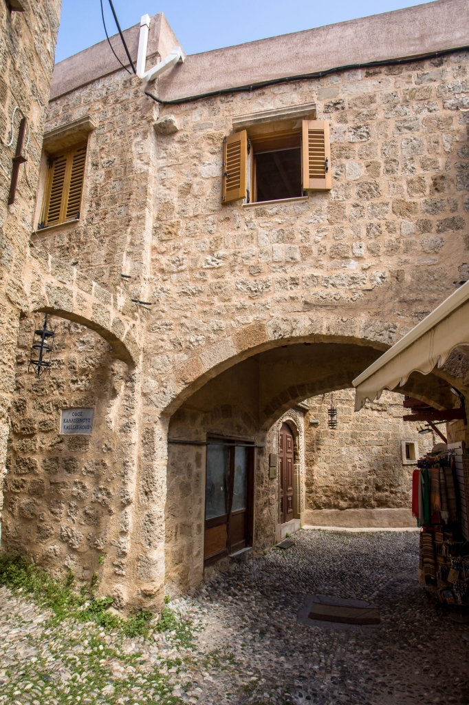 A stone building with an arched passageway, wooden shutters, and a cobblestone street in a historic Mediterranean town. [AI-generated text]