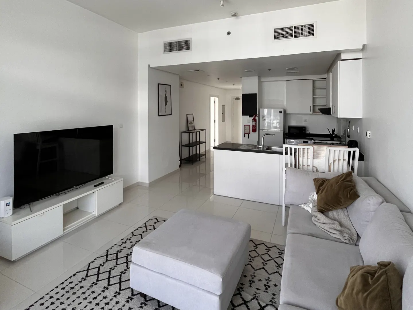 A bright, modern apartment interior with white walls, a black and white rug, and a gray sofa. A kitchen area is visible in the background.
