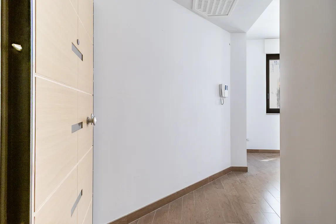 A bright hallway with wood floors and white walls. An open light wood door is on the left. A window is visible at the end of the hall.