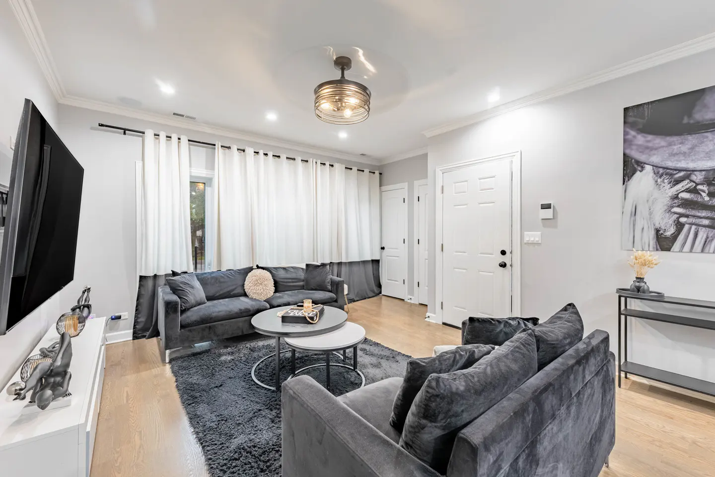 Living room with gray sofas, a dark rug, and light wood floors. White curtains cover a window. A black and white art piece hangs on the wall.