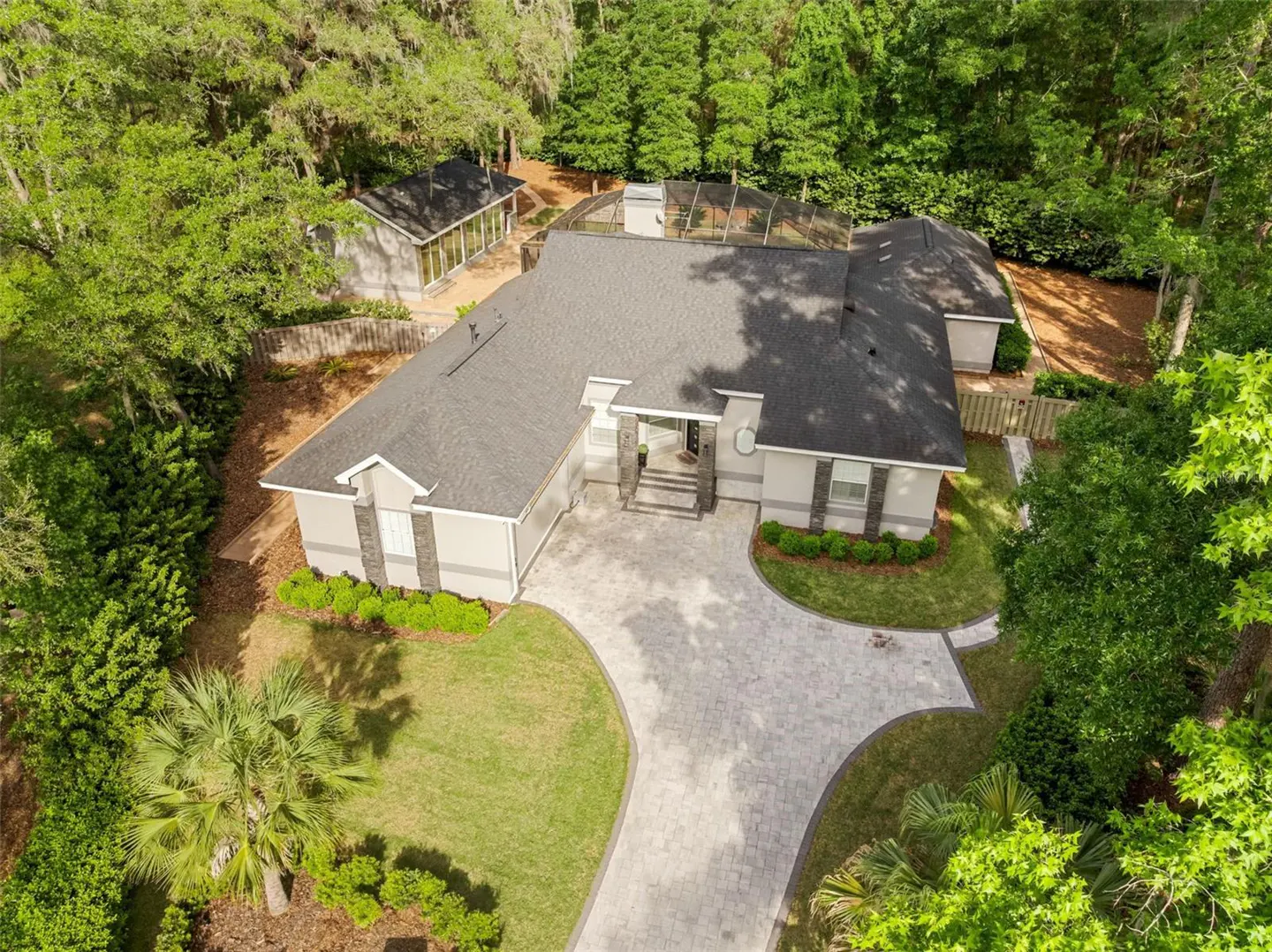 Aerial view of a one-story beige house with a gray roof, surrounded by green trees and a gray brick driveway.