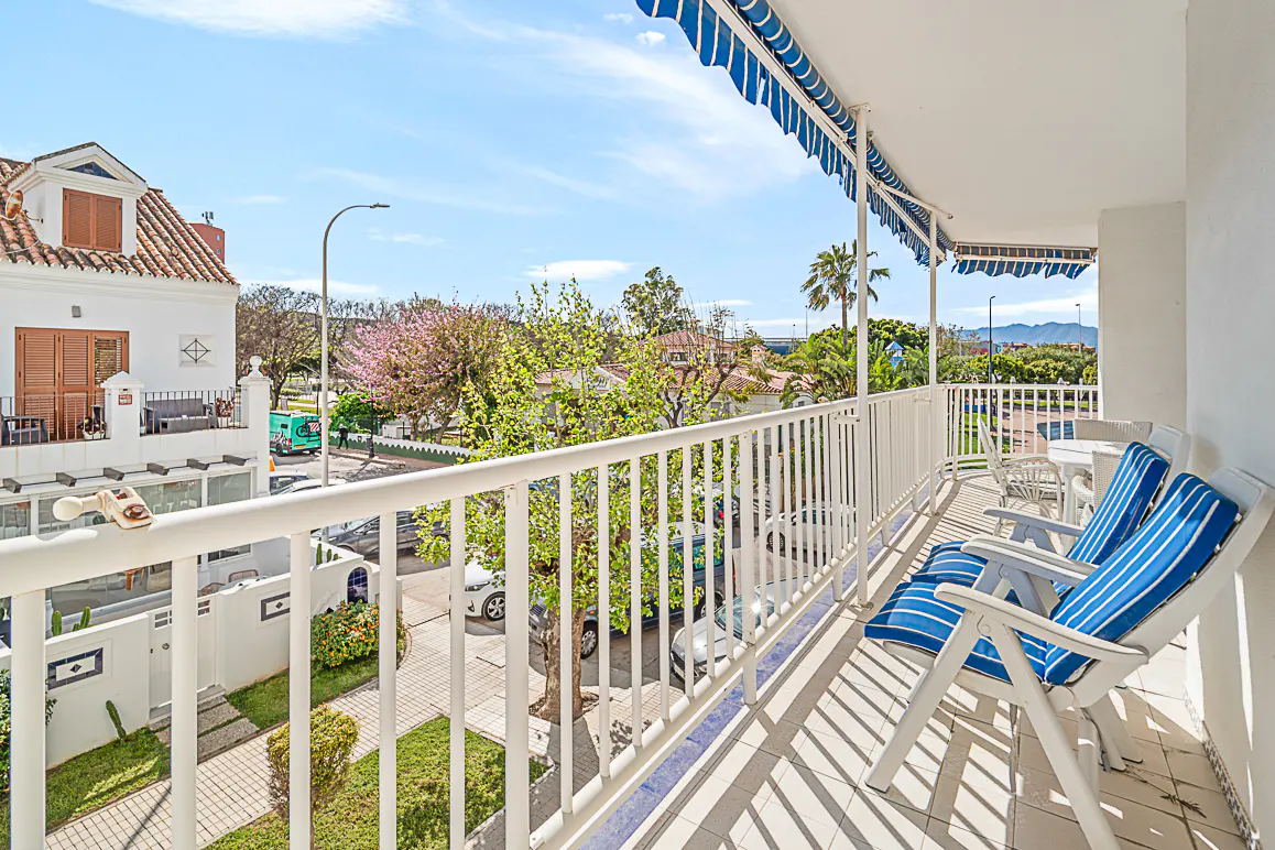 Balcony view with blue striped chairs, white railings, and a blue and white striped awning. Trees and buildings are visible in the background.