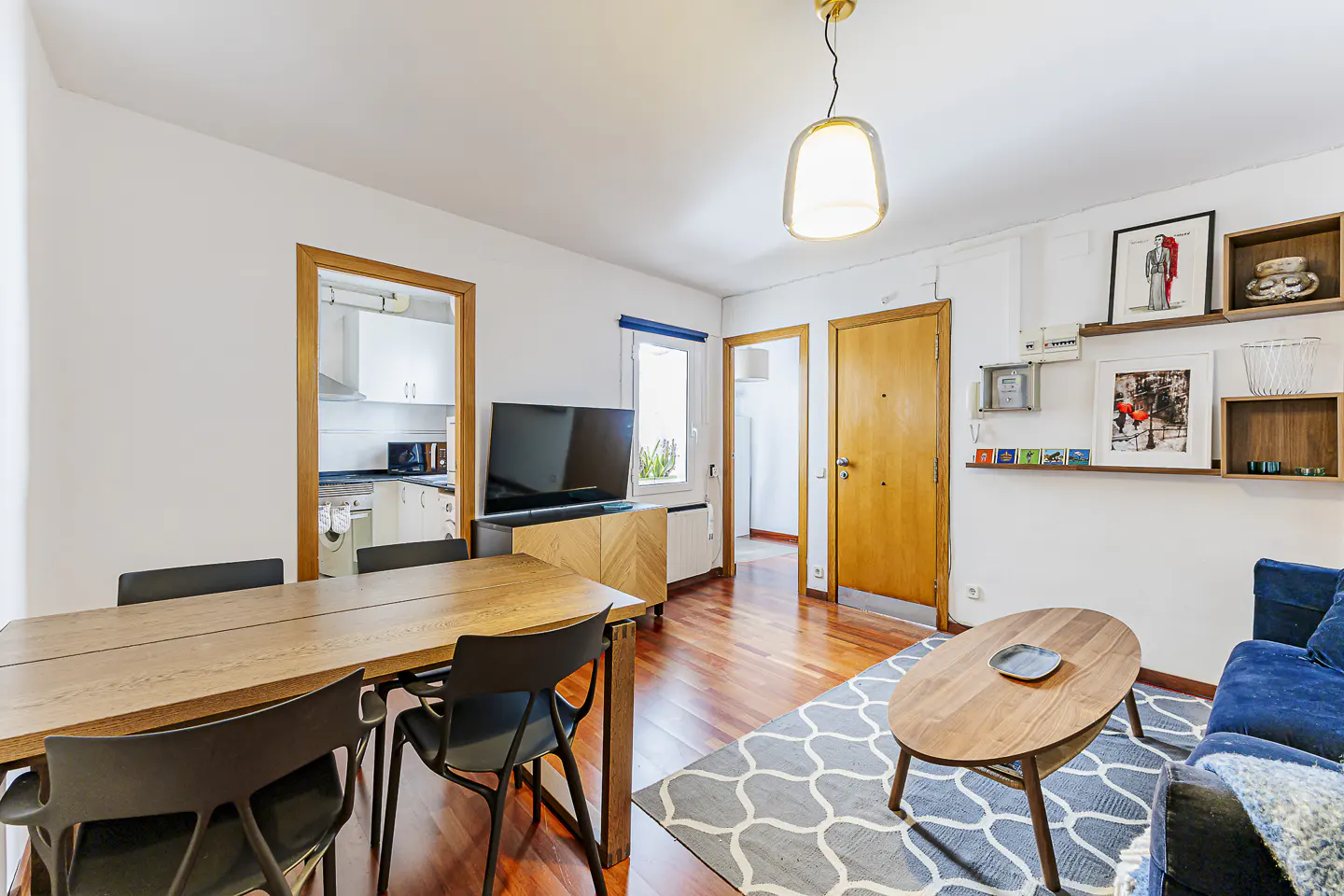 Bright living room with wood floors, a dining table with black chairs, a blue sofa, and a patterned rug. Shelves with art decorate the wall.