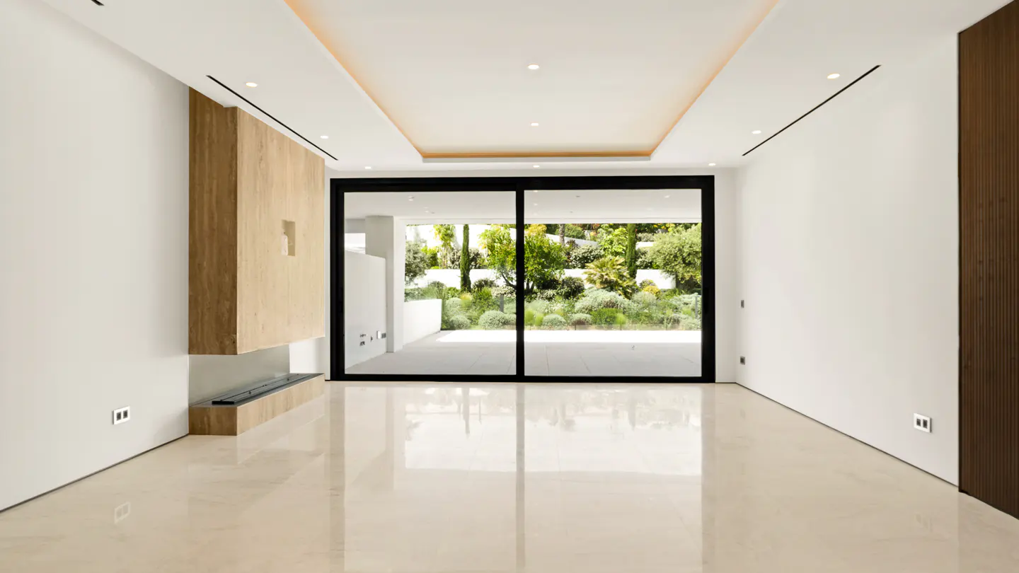 Bright, modern living room with white walls, light marble floors, and a black-framed sliding glass door to a lush green garden. A wooden fireplace adds warmth.