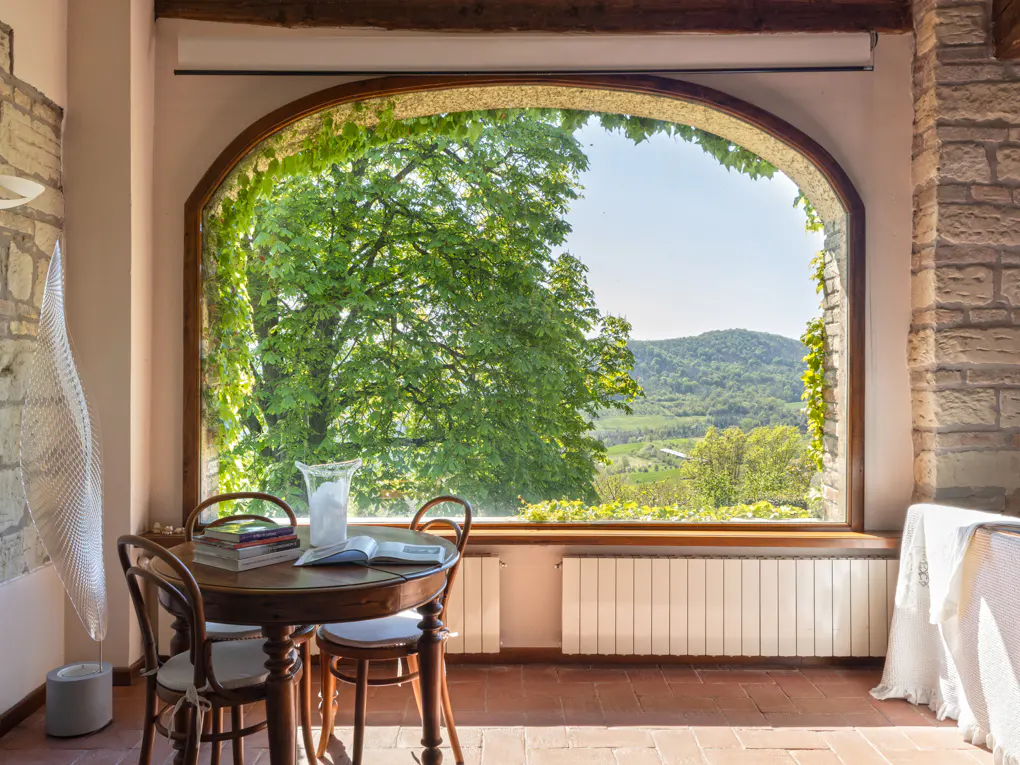 A sunlit room with a round table, chairs, and a large arched window framing a green landscape.