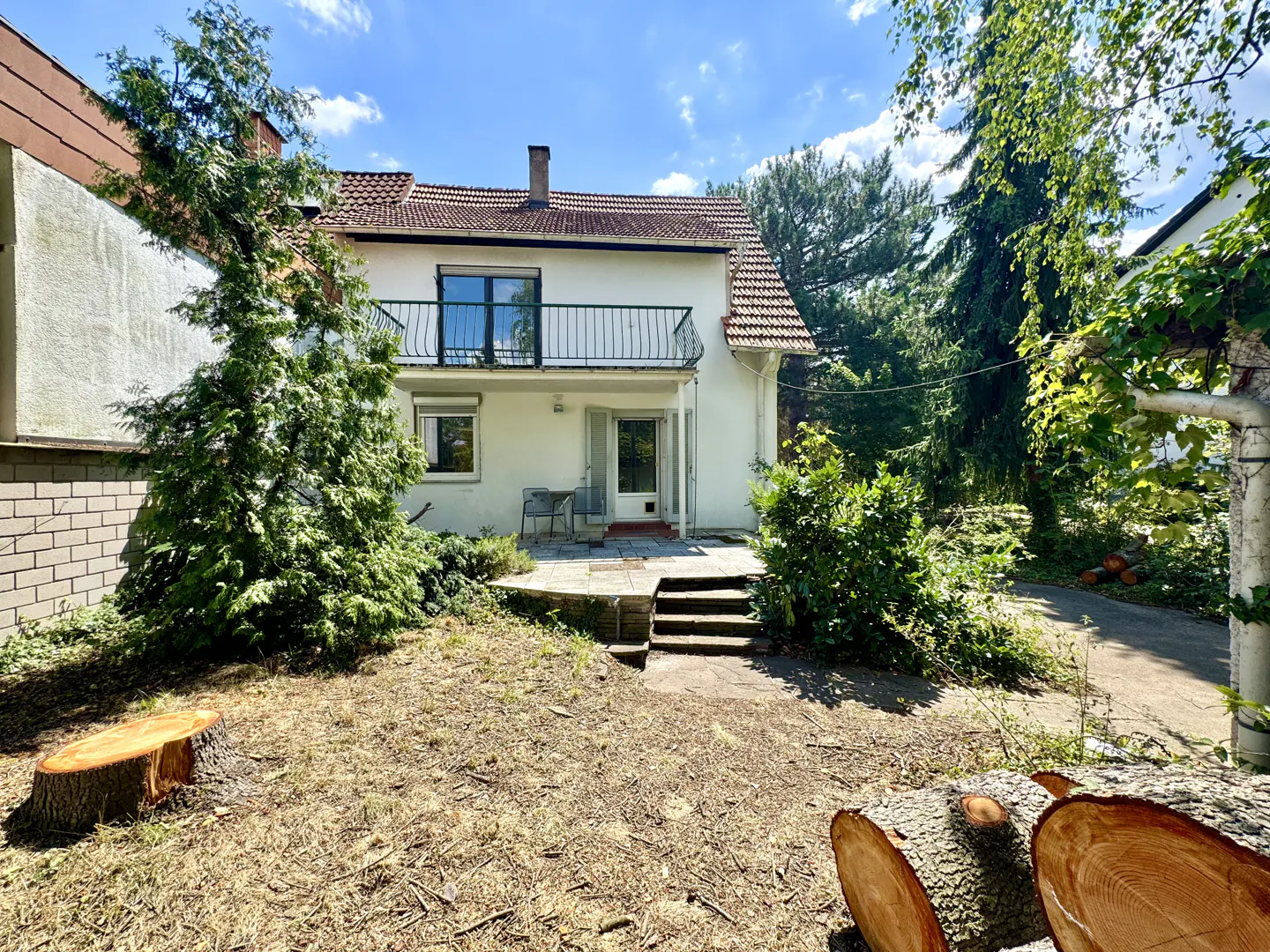 Backyard view of a two-story white house with a red tile roof, balcony, and patio. Tree stumps and logs are in the yard.