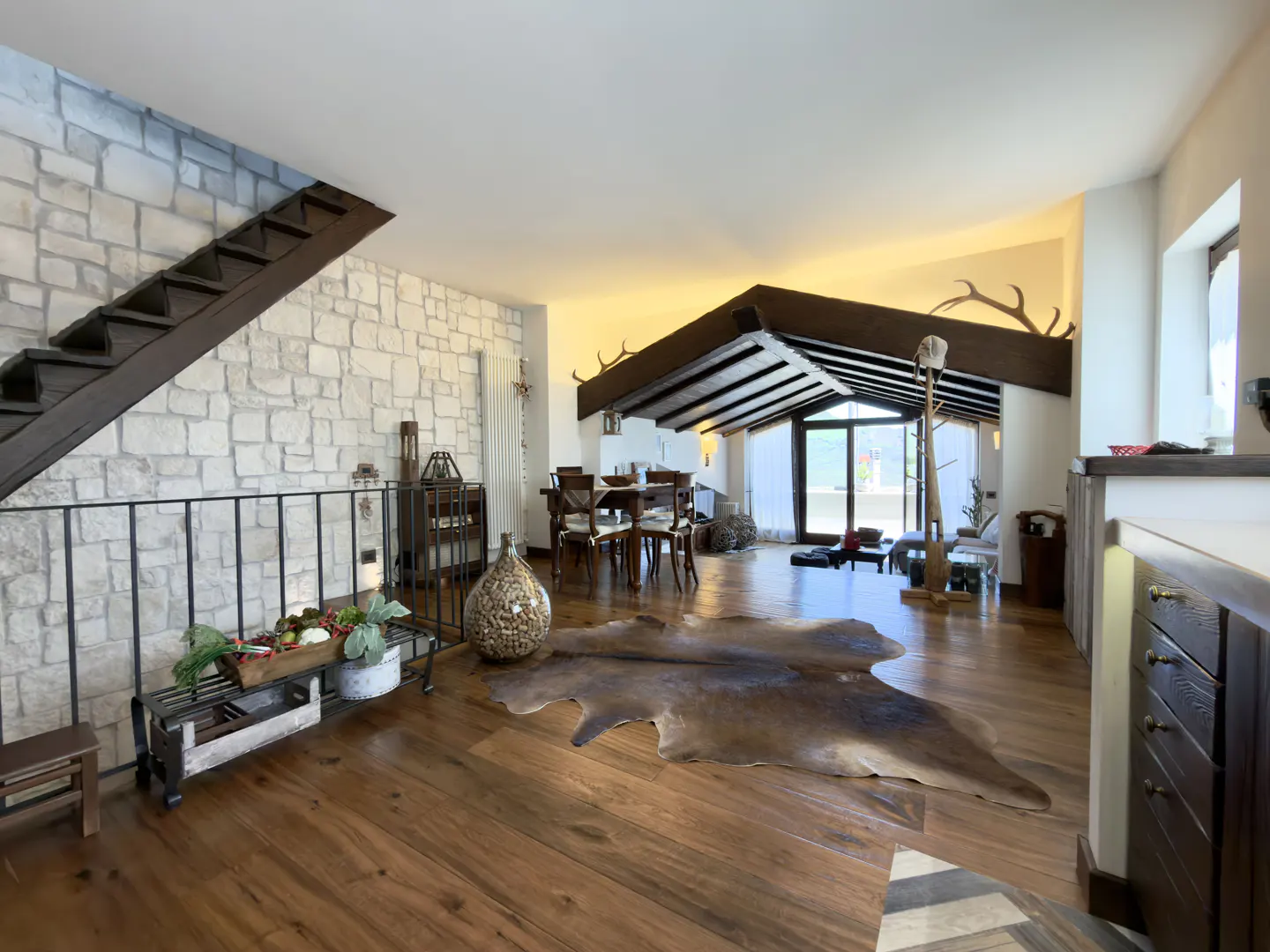 Open-concept living space with wood floors, stone wall, and dark wood beams. A cowhide rug sits in front of a dining table.