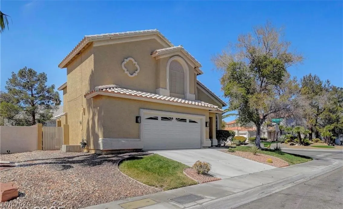 Two-story tan stucco suburban home with a white garage door, tile roof, and desert landscaping under a clear blue sky.