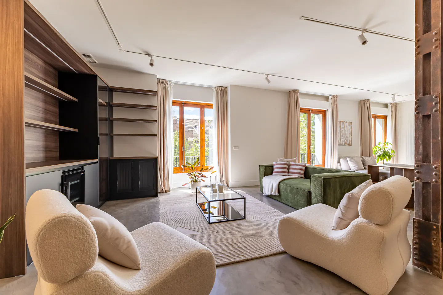 Bright living room with beige chairs, green sofa, and wood-framed windows. Built-in shelves and a glass coffee table add to the modern design.