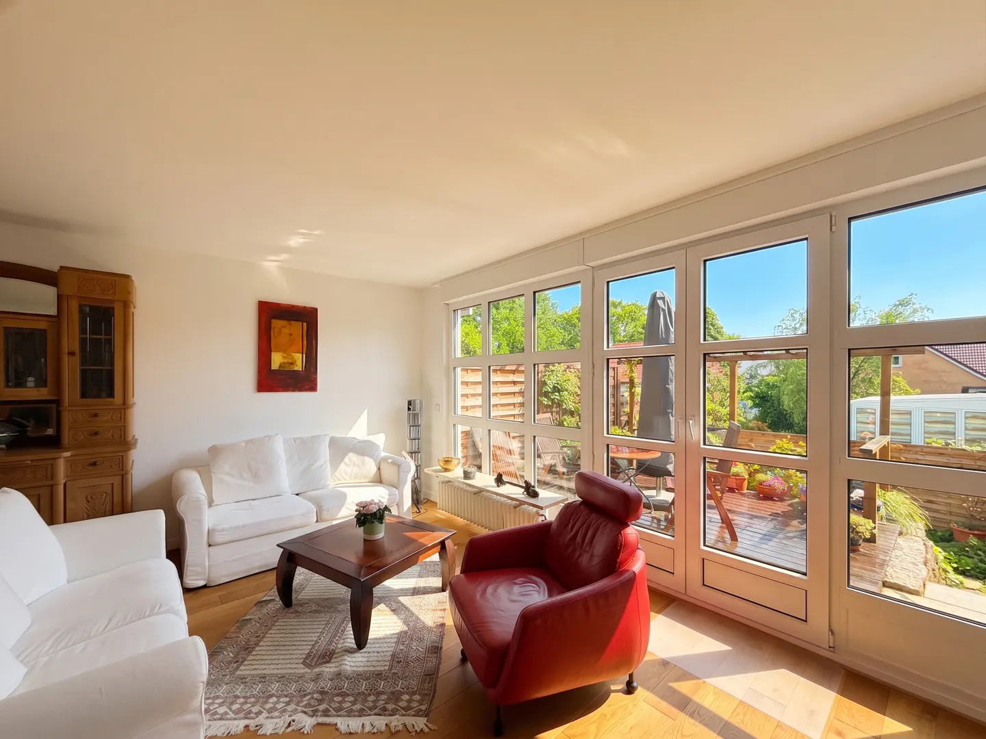 Bright living room with white sofas, red chair, wood floors, and large windows overlooking a sunny patio.