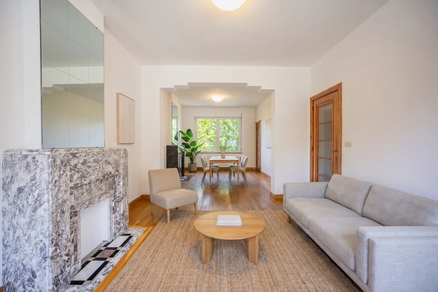 Living room with a marble fireplace, beige sofa, round table, and dining area in the background.