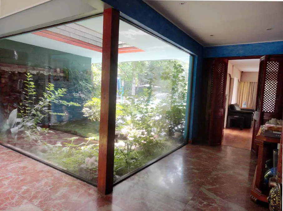 Interior view of a room with a large glass window overlooking a lush garden with plants and a waterfall feature. The room has marble floors and blue walls.