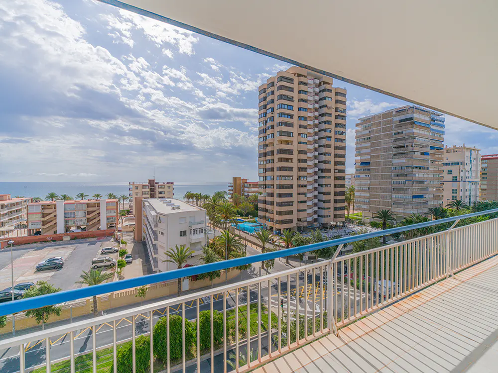 View from a balcony with white railings and blue trim, overlooking buildings, palm trees, and the ocean under a cloudy sky.