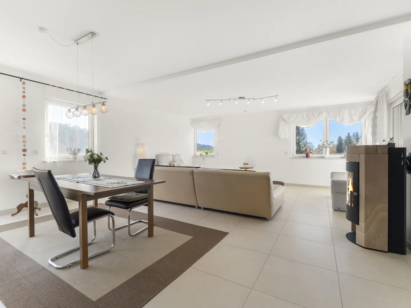 Bright, open-plan living space with dining table, beige sofa, and modern fireplace. Windows offer views of trees and blue sky.