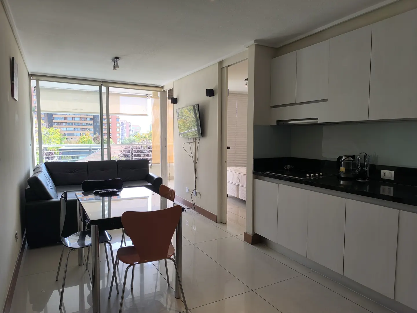 Open-concept apartment with a black leather sofa, glass table, and white kitchen cabinets. A TV is mounted on the wall.