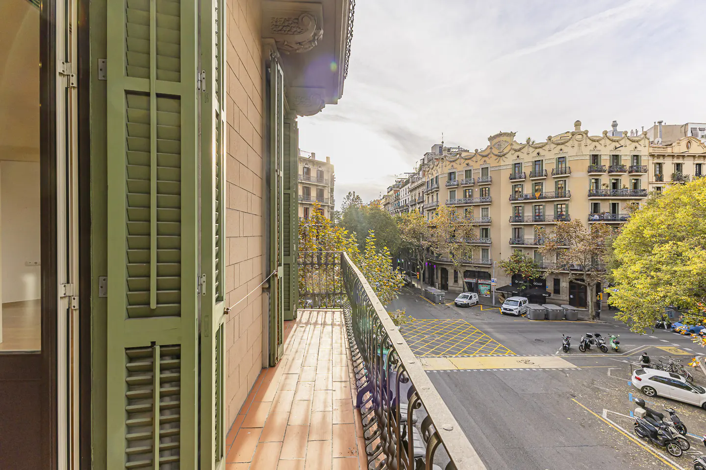 View from a balcony with green shutters overlooking a sunny city street intersection with buildings and parked vehicles.