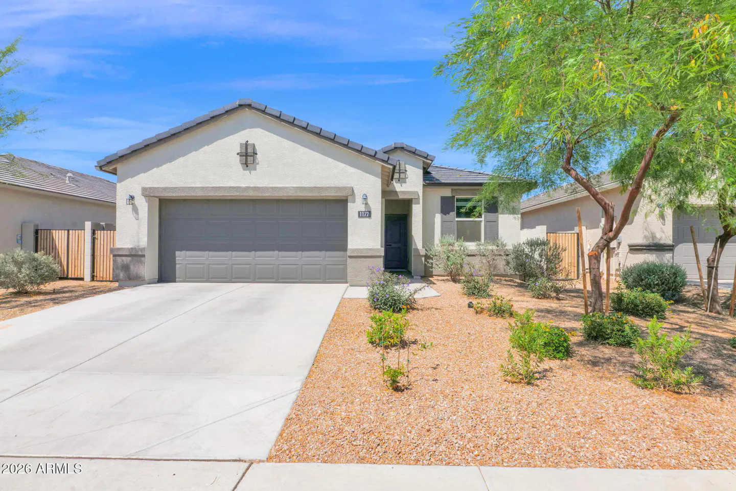 Beige single-story house with a gray garage door, gray trim, and a dark gray front door. The yard has small plants and tan gravel. Blue sky.