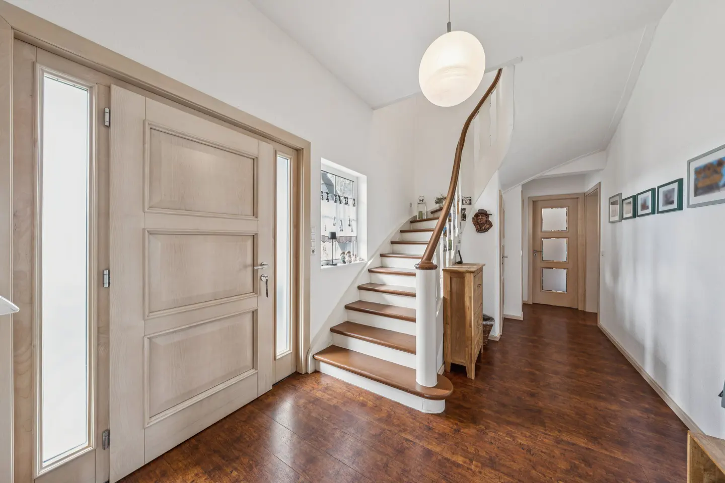 Bright foyer with wood floors, white walls, and a staircase. A front door is on the left, and pictures hang on the right wall.