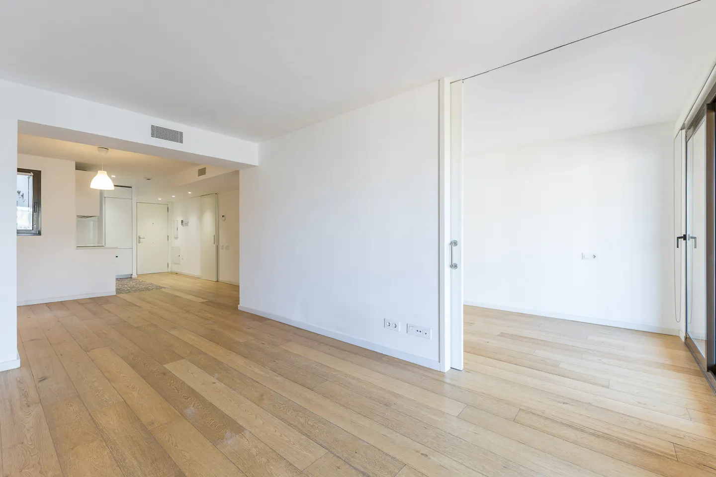 Bright, empty apartment with light wood floors and white walls. A doorway leads to another room with sliding glass doors.